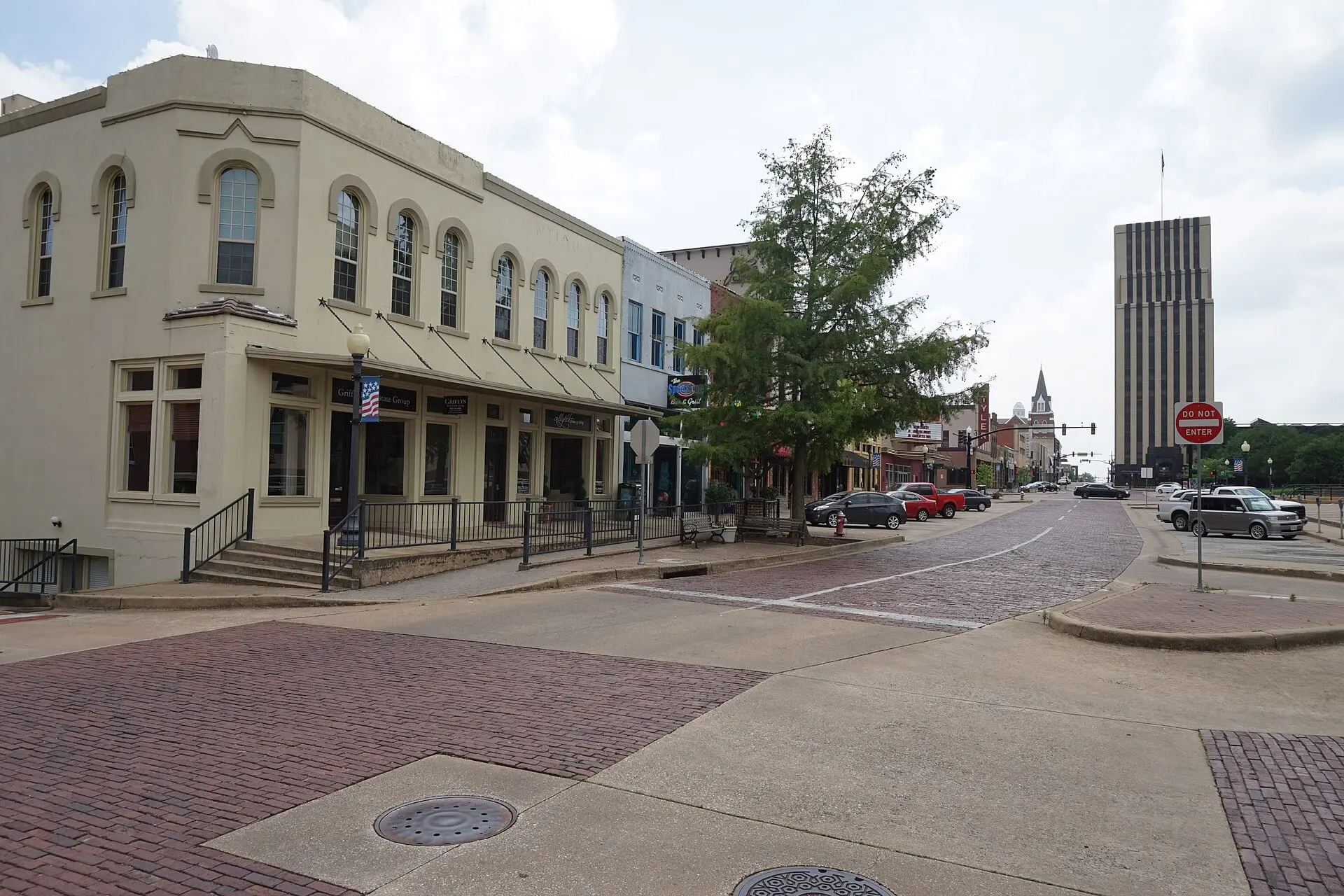 A downtown corner with brick crosswalks and brick paving, a two-story cream building at left and a tall tower in the distance.
