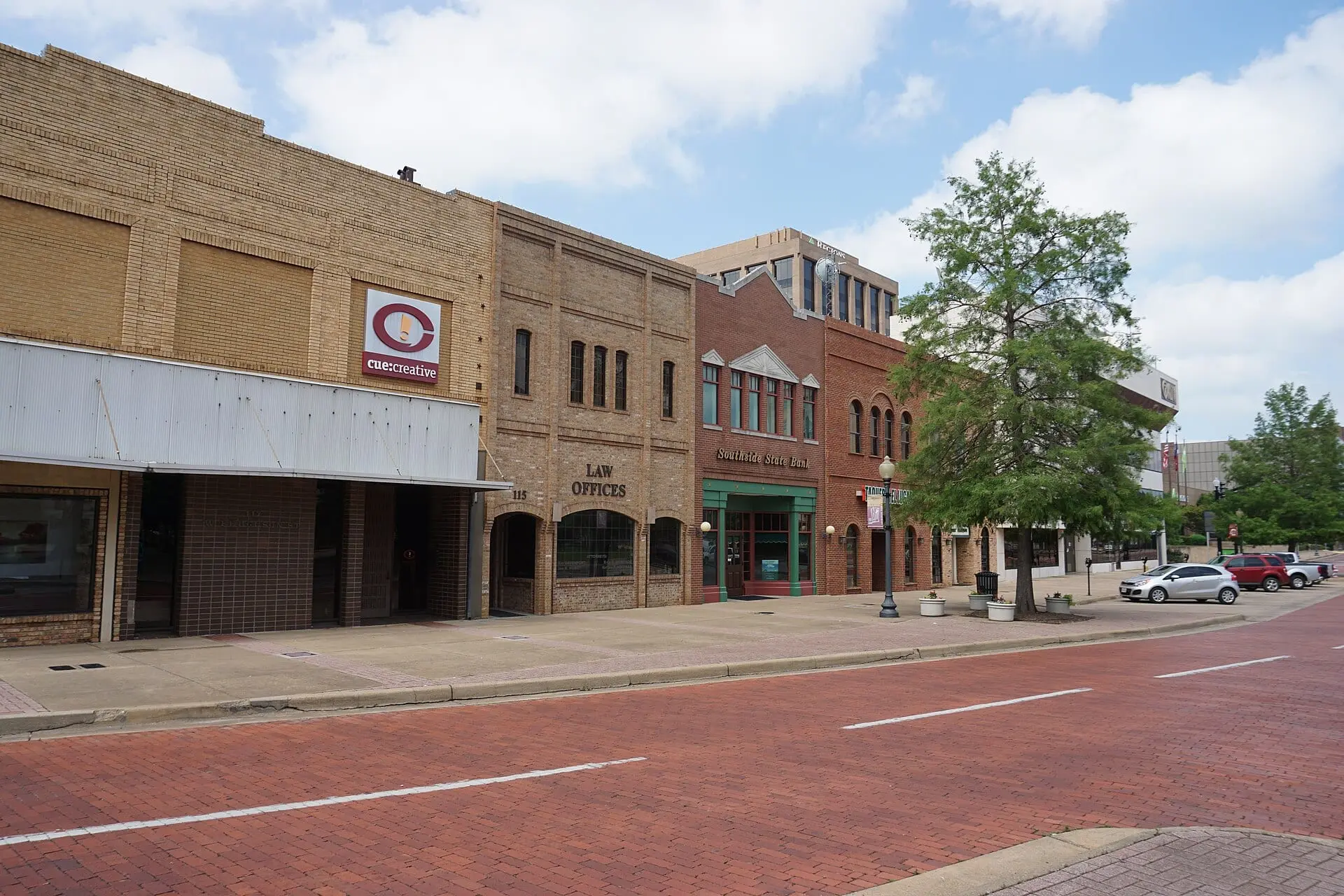 A wide street of bright red brick with white lane lines in front of a row of two-story brick storefronts.