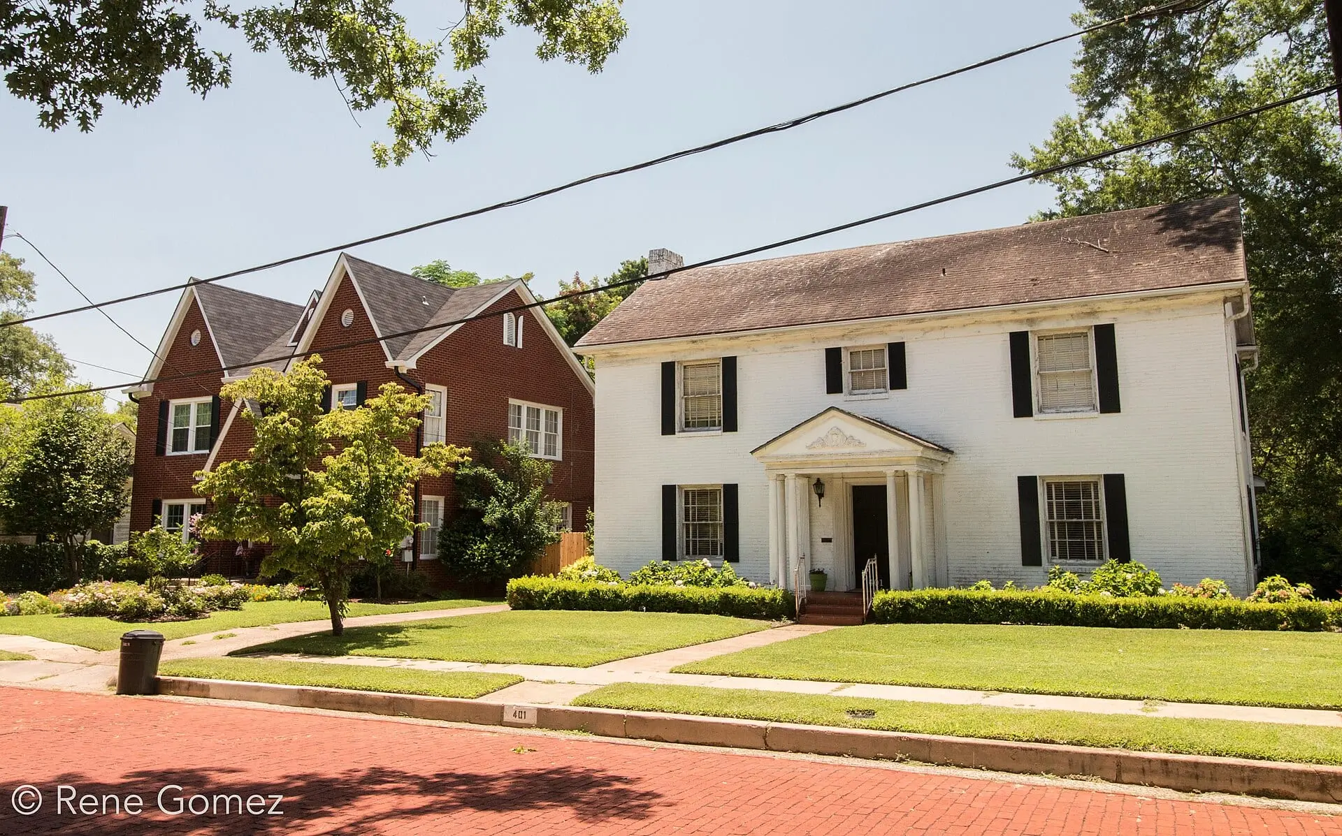 A white two-story house with black shutters and a red-brick house beside it, behind a lawn and a red-brick street.