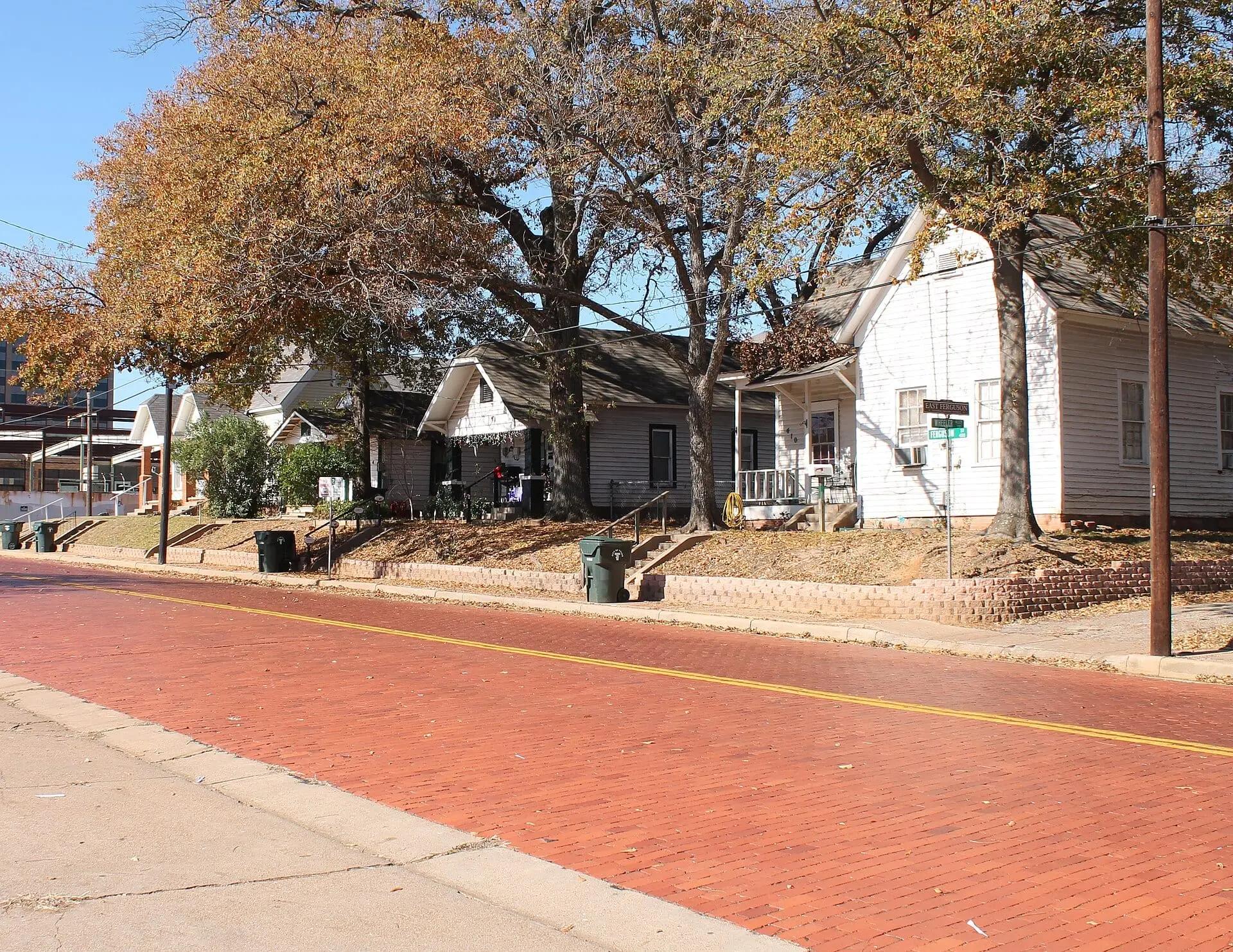 A red-brick street with a yellow center line in the foreground, small frame houses under autumn trees beyond.