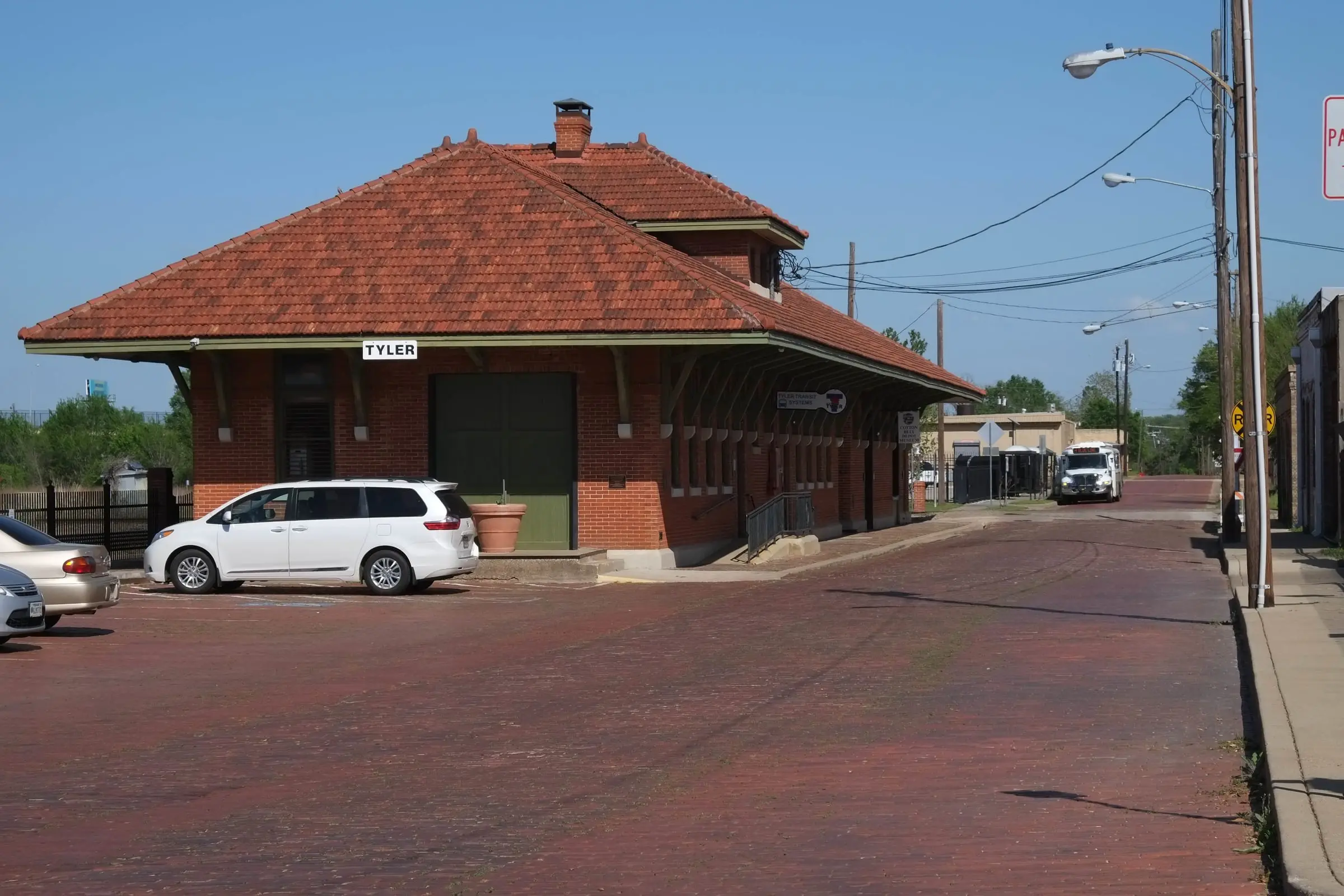 A small brick railroad depot with a red tile roof and a sign reading TYLER, a brick-paved street and parking area in front.