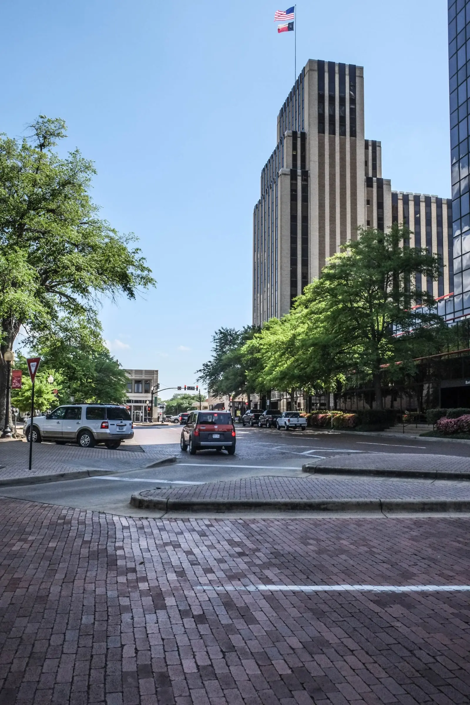 Worn brick paving in the foreground, a curb island and a street, with a tall stepped Art Deco tower above the trees.